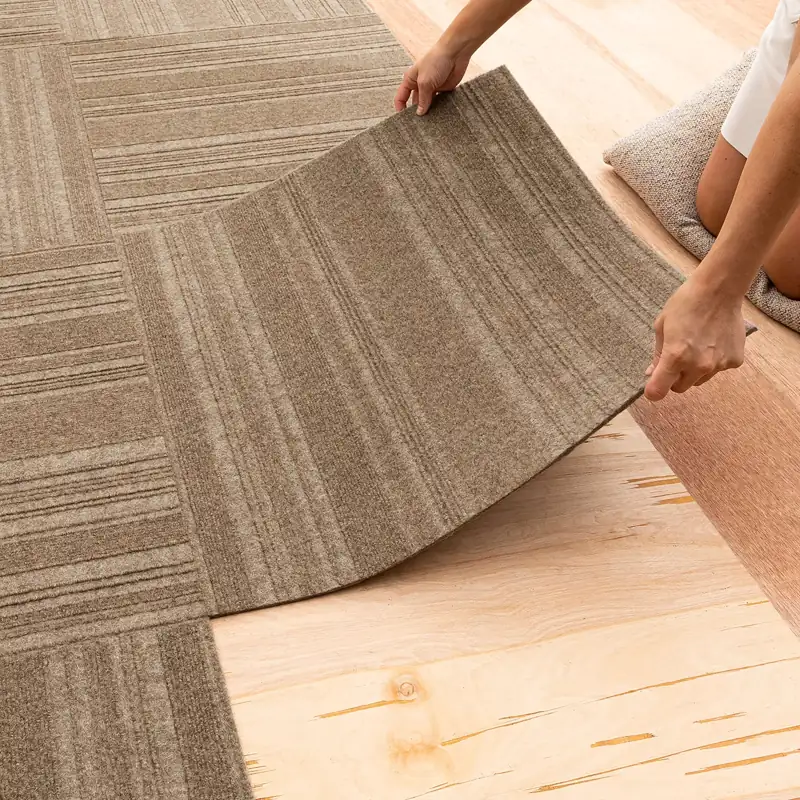 Person installing brown carpet tiles on a wood floor.